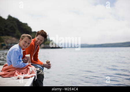 Ritratto di un sorridente ragazzo seduto con sua madre sul bordo di un pontile tenendo una bevanda al cioccolato. Foto Stock