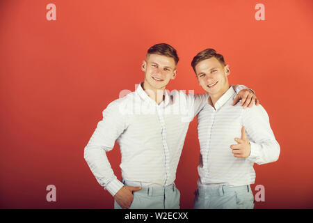 La famiglia, di fraternità e di amicizia concetto. Felice uomini avvolgente. Due fratelli sorridente e puntare il dito. I modelli in piedi su sfondo rosso. Gemelli usura Foto Stock