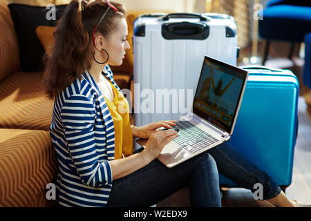 Vista laterale del moderno viaggiatore Donna Camicia a Righe seduto vicino al lettino e borse trolley a casa moderna nella soleggiata caldo giorno d'estate piano di acquisto tic Foto Stock