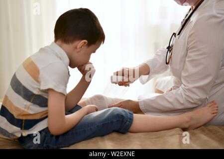 Il medico in camice bianco fornisce attentamente prima di aiuti. Mettere un cerotto sul ragazzo in gamba. Il kid pazientemente tiene il suo mento con la sua mano e osserva. Foto Stock