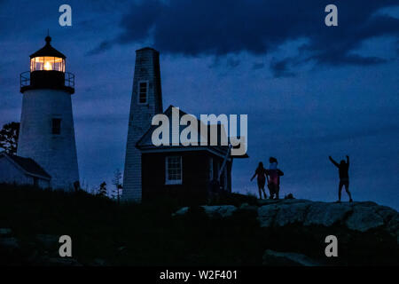Persone stagliano dal cielo al crepuscolo come essi a piedi sulla spiaggia rocciosa presso la storica Pemaquid Point Lighthouse in Bristol, Maine. Il pittoresco faro costruito lungo la costa rocciosa di Pemaquid Point è stata commissionata nel 1827 dal Presidente John Quincy Adams. Foto Stock
