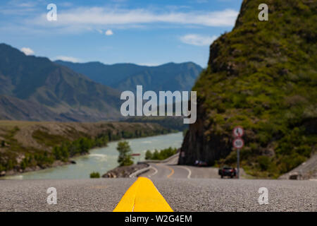 Close-up su un giallo striscia divisoria su una strada asfaltata che si protende nella montagne di Altai vicino ad una roccia con rocce e un fiume con un pittoresco terre Foto Stock