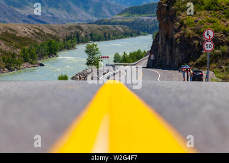 Close-up su un giallo striscia divisoria su una strada asfaltata che si protende nella montagne di Altai vicino ad una roccia con rocce e un fiume con un pittoresco terre Foto Stock