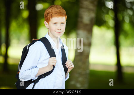 Sorridente redheaded scolaro con zaino nero e in maglia bianca di andare a scuola per il suo primo giorno di lezioni Foto Stock