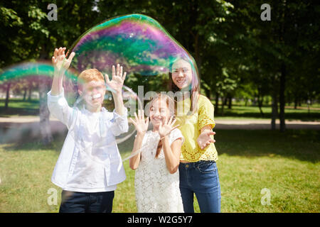 Jolly eccitato multi-etnico kids in abiti casual giocando con grandi bolle di sapone insieme mentre si divertono nel parco di estate Foto Stock