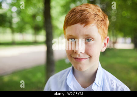 Contenuto carino redhead ragazzo in maglietta blu in piedi di green park e guardando la fotocamera Foto Stock