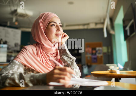 Abbastanza positivo giovane donna in hijab iscritto i suoi sogni in Diario quando si è seduti a tavola in cafe Foto Stock