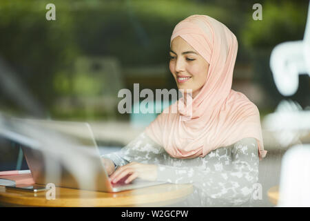 Abbastanza positivo giovane donna in rosa hijab rispondere a messaggi di posta elettronica sul computer portatile Foto Stock