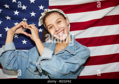 Ritratto di jolly giovane patriota d'America indossa velo in piedi contro la bandiera nazionale e unire le dita nella forma del cuore Foto Stock