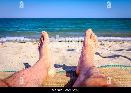 L uomo per le gambe fino a quando non viene a prendere il sole in giacente spensierato sul tappeto accanto alla linea di costa, sulla spiaggia pubblica. Foto Stock