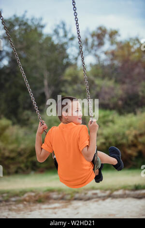 Una vista posteriore colpo di un giovane ragazzo caucasico indossando un abbigliamento casual, egli è divertirsi su altalena in un parco pubblico. Foto Stock