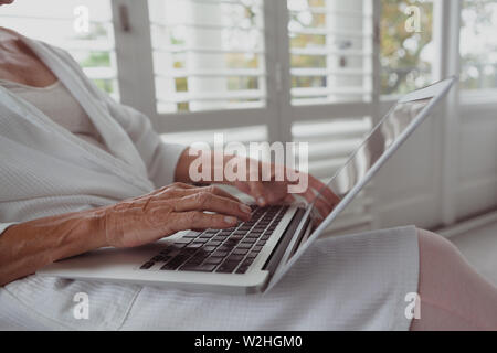 Attiva donna senior utilizzando computer portatile su una sedia in una casa confortevole Foto Stock