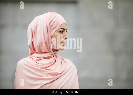 Vista laterale del contenuto introspettivo giovane donna musulmana di fede islamica di indossare il hijab rosa cercando in distanza all'aperto Foto Stock