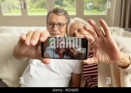 Active senior giovane seduto sul divano e prendendo selfie in una casa confortevole Foto Stock