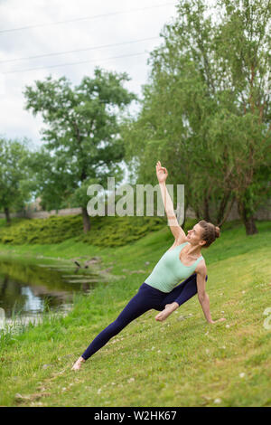 Slim bruna giovane ragazza yogi non difficile esercizi yoga sull'erba verde sullo sfondo dell'acqua. Foto Stock