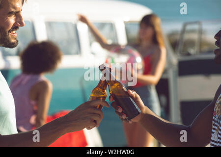 Amici maschi tostare bottiglia di birra nei pressi di camper presso la spiaggia Foto Stock