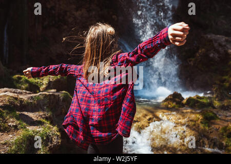 Bella felice bambina sta godendo del paesaggio di montagna, passando le mani e guardando la splendida cascata all'aperto. I suoi capelli soffia in th Foto Stock