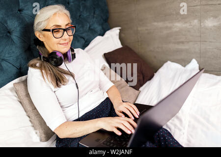 Senior donna con i capelli bianchi in occhiali che indossano le cuffie in chat sul laptop mentre posa sul letto Foto Stock