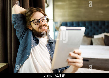 Sorridente barbuto hipster l uomo con gli occhiali seduto con tablet e leggendo qualcosa in camera da letto Foto Stock