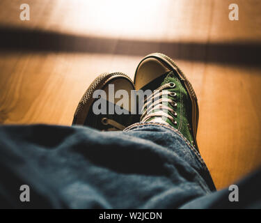 Prospettiva in prima persona su due gambe incrociate indossando sneakers retrò e jeans seduti, pavimento in legno, moody luci e ombre, look vintage Foto Stock