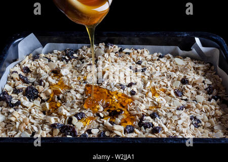 Granola in un nero vassoio da forno con la carta pergamena pronto per essere cotto al forno con miele versato su dal lato Foto Stock