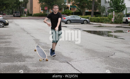 Un giovane uomo che indossa una camicia nera ed occhiali di pantaloncini verde con scarpe blu lancia un longboard giù sul terreno per ottenere una testa rullatrice start. Foto Stock