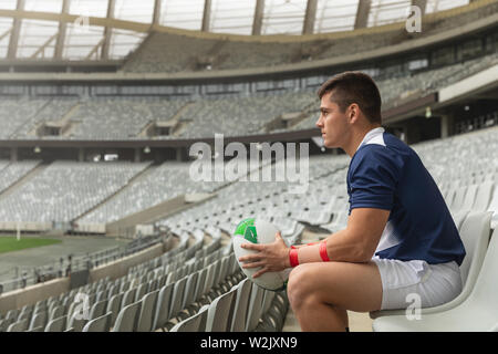 Maschio caucasico rugby giocatore seduto con palla da rugby in stadium Foto Stock