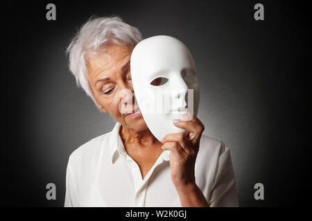 Donna anziana con i capelli bianchi nascondendo il volto triste e dietro la maschera - depressione o il concetto di personalità Foto Stock
