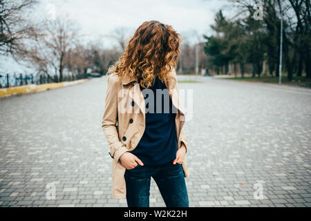 Irriconoscibile giovane donna con capelli ricci indossando il beige trench coat in piedi nel parco della città tenendo le mani nelle tasche. Foto Stock