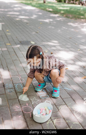 Carino il toddler ragazza seduta e le forme di disegno su asfalto nel parco. Bambino azienda chalk. Foto Stock