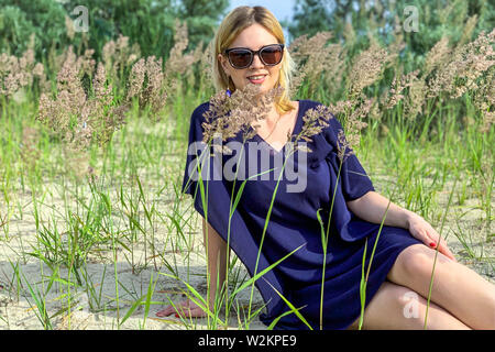 Bella donna bionda in vestito blu e occhiali da sole in posa mentre è seduto a terra tra l'erba a prato estivo, guardando la telecamera con sorriso toothy. Foto Stock