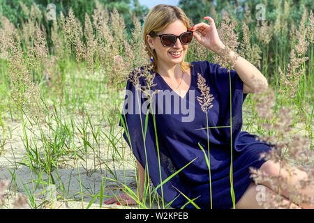 Bella donna bionda in vestito blu e occhiali da sole in posa mentre è seduto a terra tra l'erba a prato estivo, tenendo i suoi occhiali e cercando wi Foto Stock