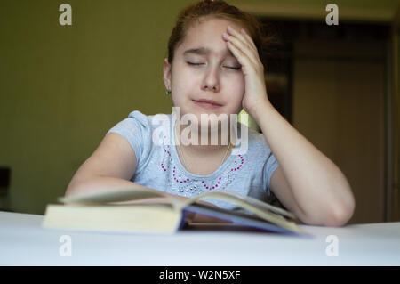 Torturato ragazza bambino legge un libro a tavola e lei è molto stanco e vuole dormire, sbadigli, si addormenta durante la lettura di storie e di lezioni Foto Stock