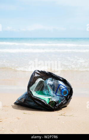 Vista dettagliata del grigio di un sacchetto di plastica con rifiuti raccolti sulla sabbia di una spiaggia solitaria, accanto all'acqua, con l'orizzonte in background e alcuni bl Foto Stock