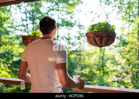 Appendere le piante in vaso con fiori viola foglie in primavera con uomo in piedi sul portico di casa in legno di mattina cottage di cabina Foto Stock