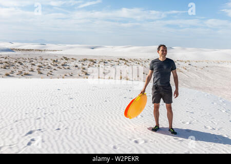 Uomo felice sulla sabbia in sabbie bianche dune monumento nazionale nel Nuovo Messico tenendo premuto il disco colorato slitta di scorrimento giù per le colline Foto Stock