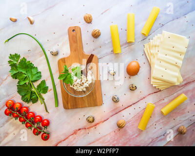 Cannoli di pasta e lasagne concetto di cucina a vista. Vista superiore degli ingredienti su un piano di marmo con un tagliere di legno: pomodori verdi, uova, formaggio. Foto Stock