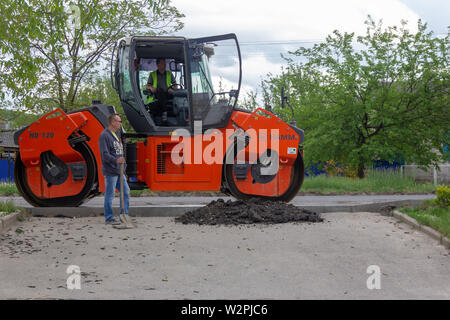 Kamennomostsky, Russia - 10 Maggio 2019: un uomo con una pala nelle sue mani è di circa asfalto rullo compattatore Hamm Foto Stock