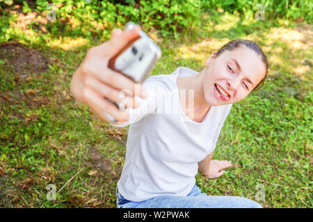 Ragazza giovane prendere selfie dalle mani di un telefono che mostra la linguetta e divertente faccia seduti sul parco o giardino sfondo. Ritratto di giovane donna attraente ma Foto Stock