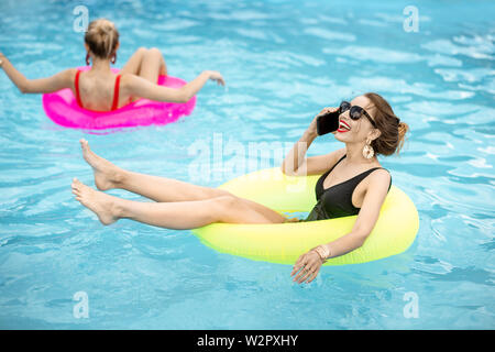 Giovane donna di parlare con il telefono durante il nuoto con anello gonfiabile in acqua piscina all'aperto durante la stagione estiva Foto Stock