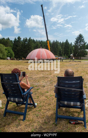 Benneckenstein, Germania. 10 Luglio, 2019. Una gru solleva il tetto di un ex cupola radar sui terreni della Ostdeutsche Fahrzeug- und Technikmuseum per sollevarlo sulle pareti dell'impianto. In primo piano, gli appassionati di tecnologia sit in sedie a sdraio. La cupola radar si fermò sul Brocken fino agli inizi degli anni novanta e schermato militari sensibili degli impianti tecnici. Il 13 luglio 2019 e 14 Luglio 2019 4 giorni di tecnologia avrà luogo nel Museo della Tecnologia. Quindi la cupola sarà visibile. Credito: Klaus-Dietmar Gabbert/dpa-Zentralbild/ZB/dpa/Alamy Live News Foto Stock