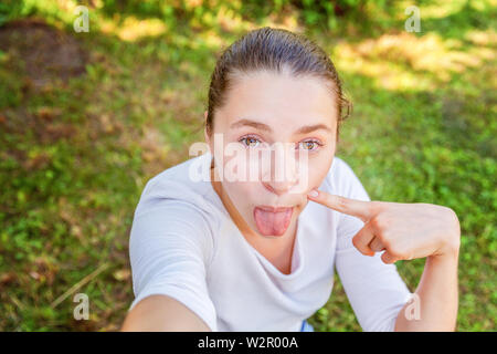 Ragazza giovane prendere selfie dalle mani di un telefono che mostra la linguetta e divertente faccia seduti sul parco o giardino sfondo. Ritratto di giovane donna attraente ma Foto Stock