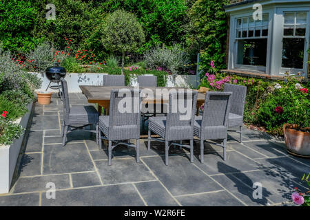 Tavolo in legno grigio e sedie di vimini su un moderno patio con un round bar-b-que in background. Splendidi fiori che circonda la pietra grigia patio. Foto Stock