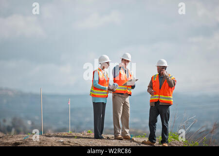 Tre ingegneri indossando alta visibilità giubbotti hardhats e discutere i progetti di un edificio su un sito in costruzione. Foto Stock