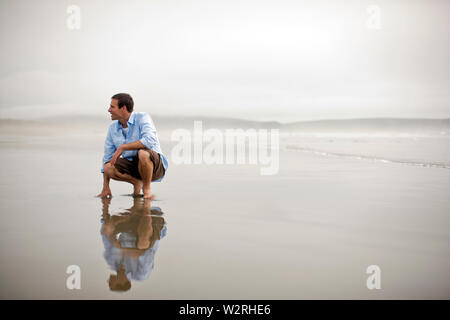 Allegro uomo accovacciato su una spiaggia con la bassa marea. Foto Stock