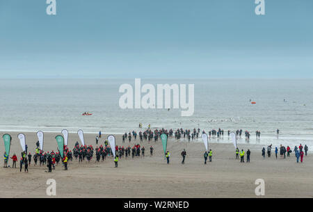 Sotto le nuvole scure concorrenti nel 2019 Barry Island sprint triathlon corrono attraverso la spiaggia di sabbia e nel mare all'inizio della gara. Foto Stock