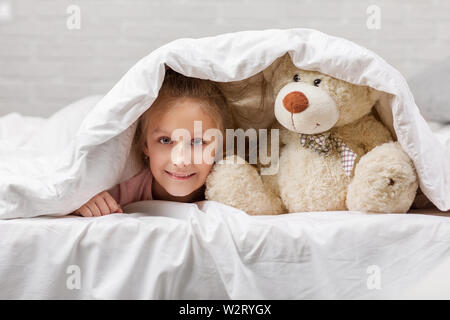 Adorabile bambina ragazza che gioca con Teddy bear nel letto nella mattina. bambino con un orsacchiotto sotto i coperchi Foto Stock