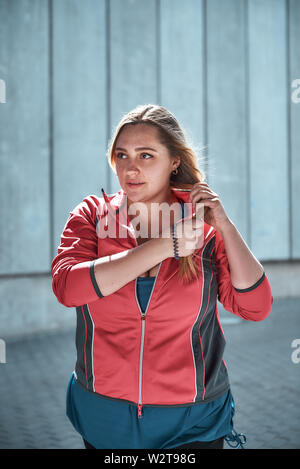 Donna sana. Foto verticale di una donna di dimensioni più belle in abiti sportivi che regola i capelli lunghi e si guarda lontano mentre si sta in piedi all'aperto. Femmina sovrappeso. Concetto di sport Foto Stock