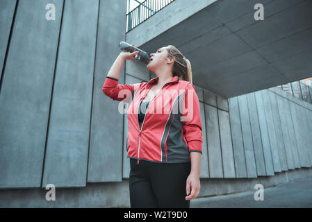 Così assetato. Giovani attraenti dimensioni plus donna in abbigliamento sportivo acqua potabile mentre facendo esercizi all'esterno. Perdita di peso. Sport concept Foto Stock