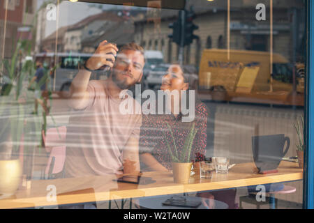 Due simpatici amici di fotografare sé, con una fotocamera del telefono - selfie. Shot attraverso la finestra con le riflessioni di strada. Foto Stock